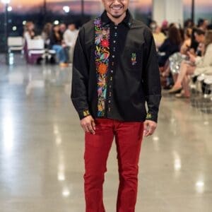Smiling male model walking a fashion runway wearing a black long-sleeve button-front guayabera shirt with a wide vertical band of multicolor hand-embroidered Otomi hummingbirds, daisy flowers, and botanical motifs along the front placket, paired with red slim-fit trousers and tan Chelsea boots