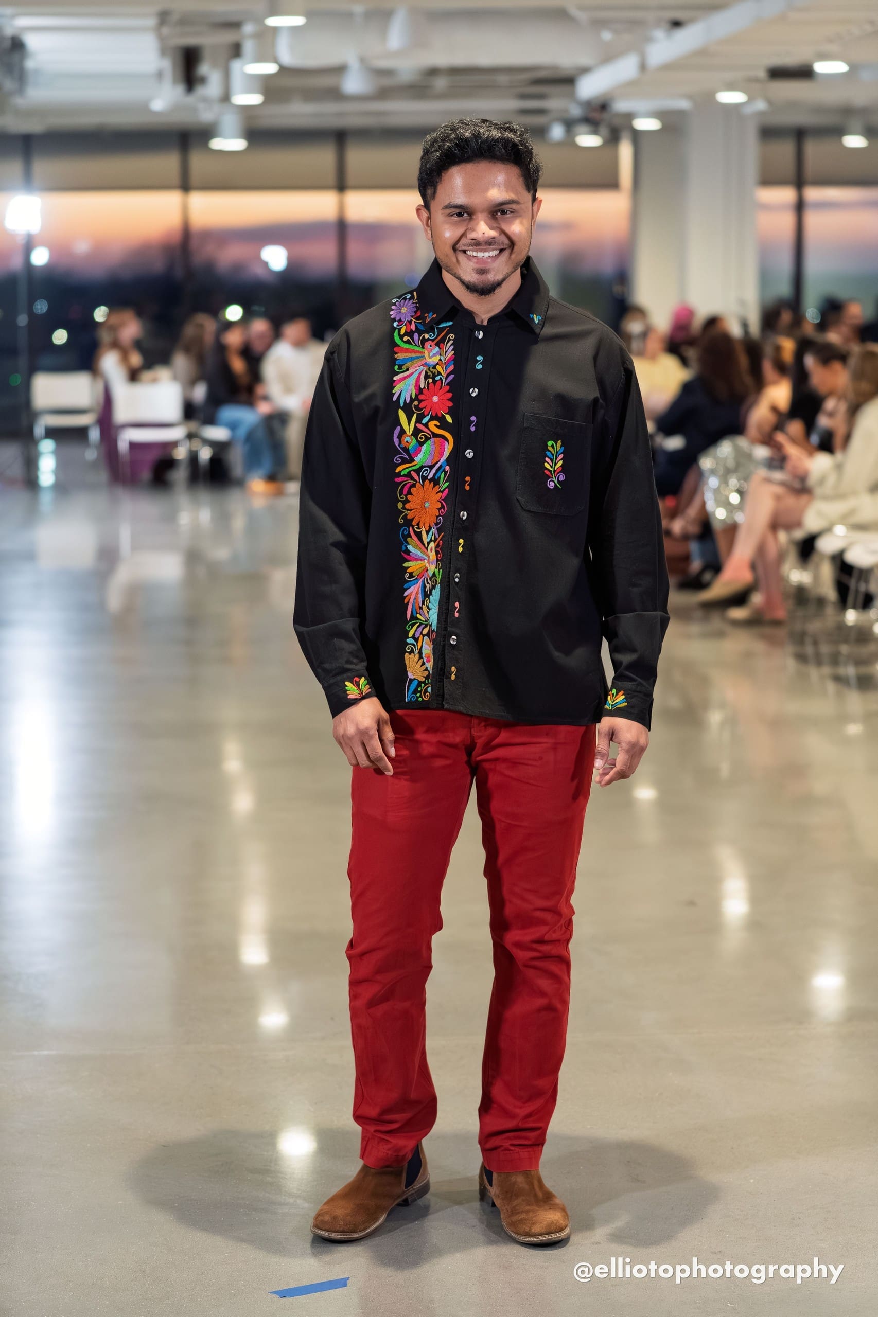 Smiling male model walking a fashion runway wearing a black long-sleeve button-front guayabera shirt with a wide vertical band of multicolor hand-embroidered Otomi hummingbirds, daisy flowers, and botanical motifs along the front placket, paired with red slim-fit trousers and tan Chelsea boots