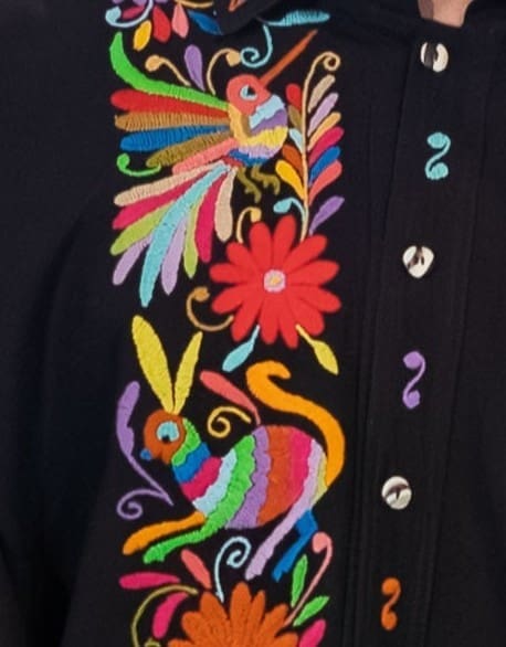 Extreme close-up of the upper front placket of a black men's guayabera shirt showing hand-embroidered multicolor Otomi hummingbird with rainbow plumes, a red daisy flower, a colorful peacock with long tail, and botanical scrolls in pink, green, teal, and orange on black cotton