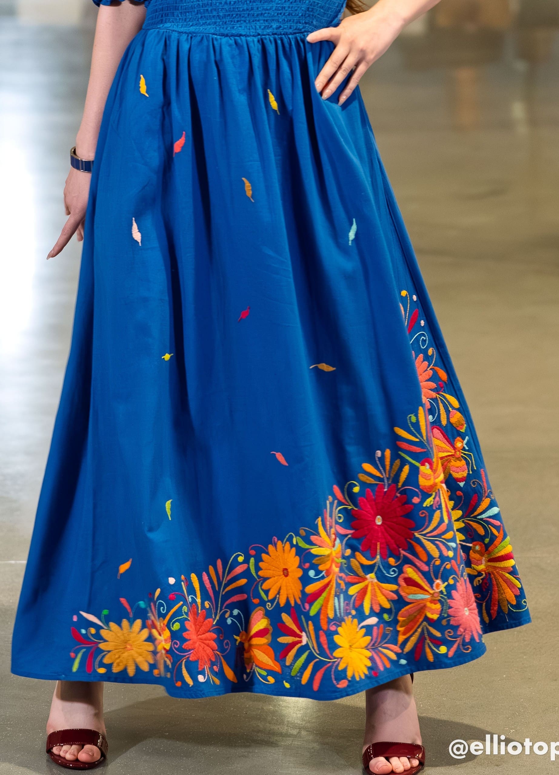 Close-up of the skirt of a cobalt blue maxi dress showing scattered multicolor hand-embroidered feather motifs across the body and a dense multicolor Otomi floral border of orange, red, and yellow daisy flowers at the hem, with a model's hand visible