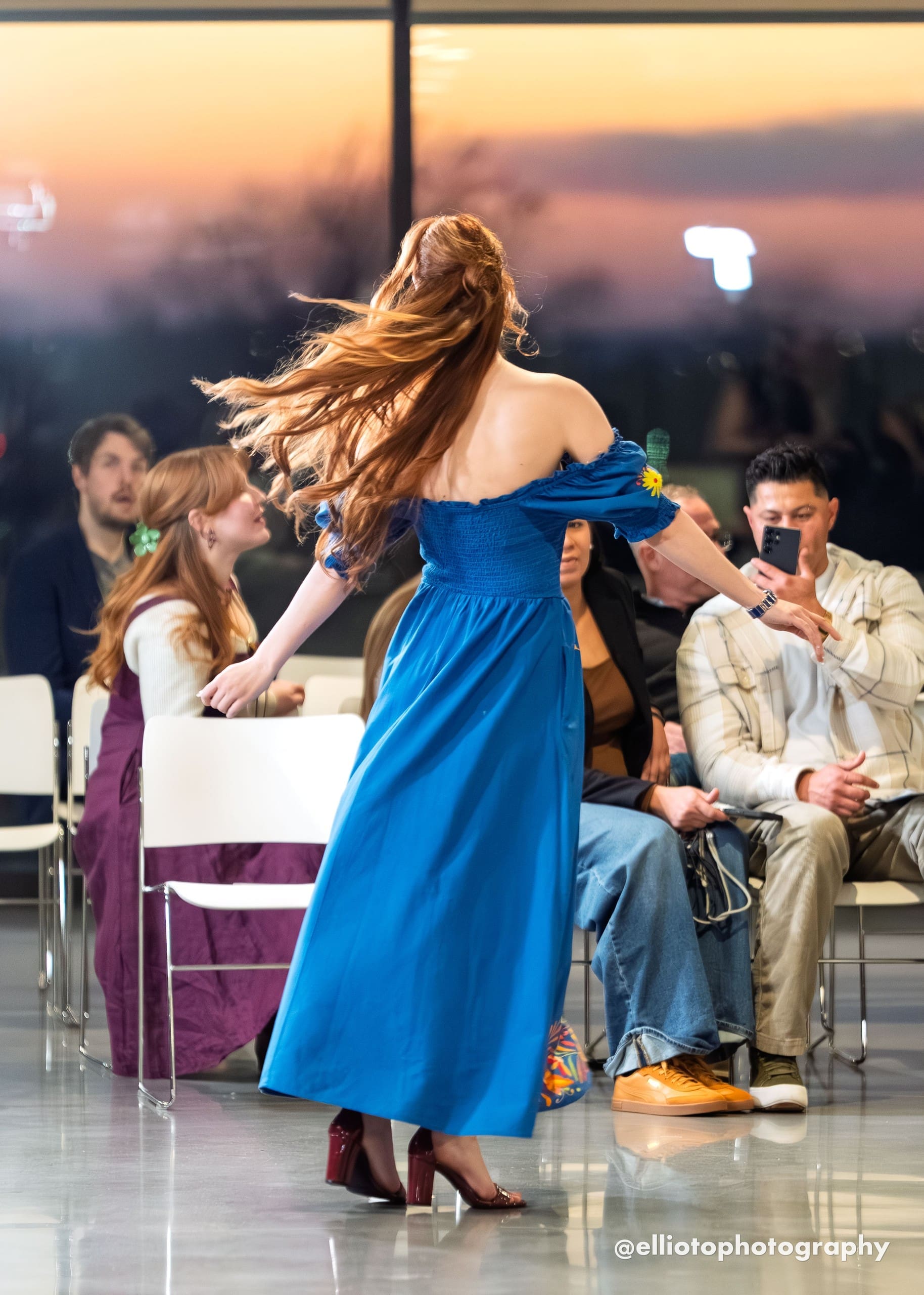 Back view of a female model mid-spin on a fashion runway wearing a cobalt blue off-shoulder smocked maxi dress, showing the plain blue back of the skirt with the multicolor Otomi embroidered border visible at the hem, hair flowing in motion