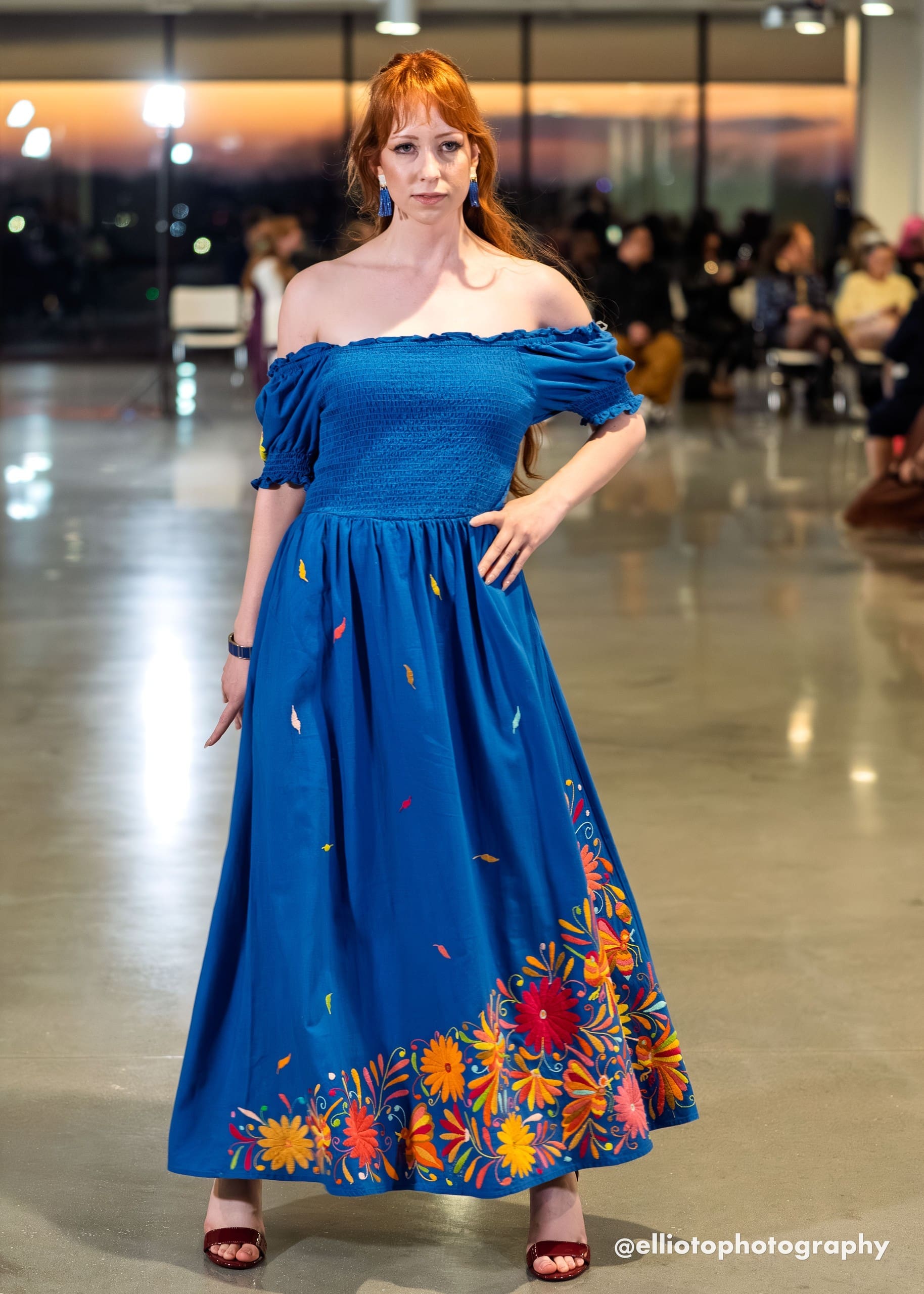 Female model on a fashion runway wearing a cobalt blue off-shoulder smocked maxi dress with multicolor hand-embroidered Otomi daisy flowers, birds, and botanical motifs along the full hem border, with scattered feather motifs on the skirt