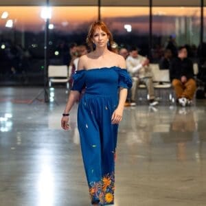 Female model walking a fashion runway wearing a cobalt blue off-shoulder smocked maxi dress with scattered multicolor hand-embroidered feather motifs on the skirt body and a dense multicolor Otomi floral border at the hem