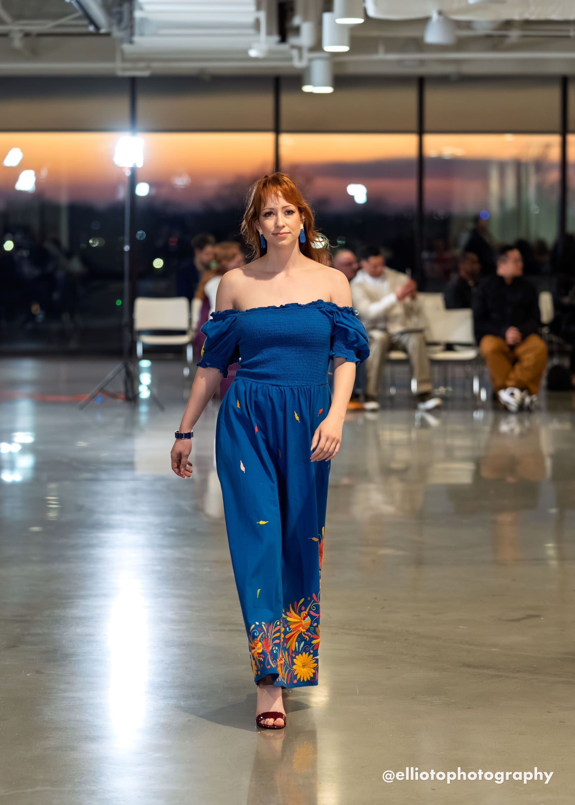 Female model walking a fashion runway wearing a cobalt blue off-shoulder smocked maxi dress with scattered multicolor hand-embroidered feather motifs on the skirt body and a dense multicolor Otomi floral border at the hem