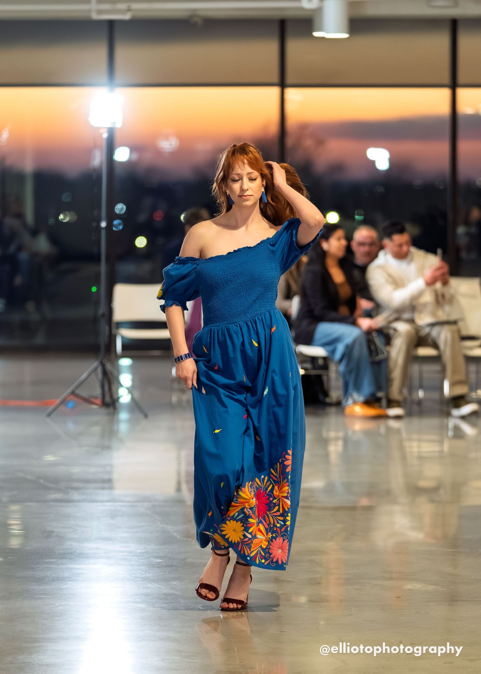 Female model walking a fashion runway wearing a cobalt blue off-shoulder smocked maxi dress with multicolor hand-embroidered Otomi daisy flowers and botanical motifs along the hem border and scattered feather motifs on the skirt, sunset backdrop visible