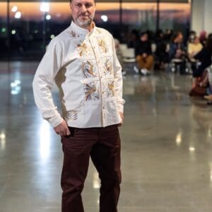 Mature male model standing on a fashion runway wearing a cream long-sleeve guayabera-style jacket shirt with hand-embroidered blue, gold, and rust Otomi bird and botanical motifs in paneled sections across the front, paired with dark brown trousers