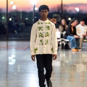 Young male model walking a fashion runway wearing a cream long-sleeve guayabera-style jacket with hand-embroidered green Otomi birds, lizards, and botanical motifs in rectangular paneled sections across the front, paired with black trousers