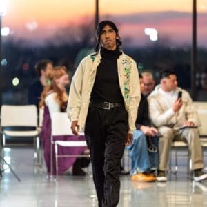 Male model walking a fashion runway wearing a cream long-sleeve zip-front jacket-style shirt with hand-embroidered teal, orange, and green Otomi birds and floral motifs along the front placket, layered over a black turtleneck with black trousers
