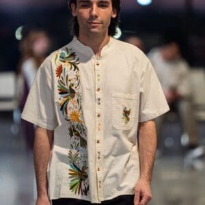 Male model wearing a cream short-sleeve mandarin-collar button-front shirt with hand-embroidered multicolor Otomi hummingbirds, daisy flowers, and botanical motifs in teal, green, brown, and orange along the front placket and chest pocket