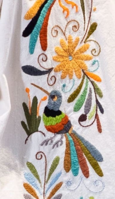 Close-up of the front placket embroidery on a cream short-sleeve guayabera shirt showing a hand-embroidered multicolor Otomi hummingbird below a golden daisy flower, with botanical leaf motifs in teal, orange, rust, olive green, and navy blue on cream cotton fabric
