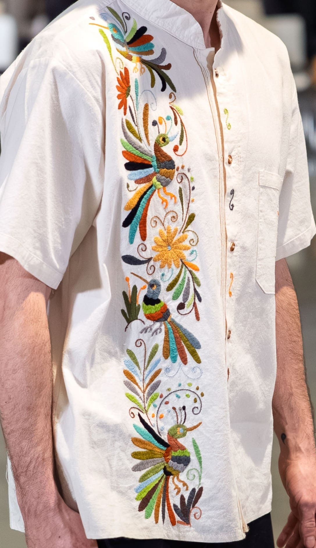 Close-up of a male model wearing a cream short-sleeve mandarin-collar guayabera shirt with a wide vertical band of multicolor hand-embroidered Otomi hummingbirds, a golden daisy flower, and botanical motifs in teal, orange, rust, olive, and black along the front placket