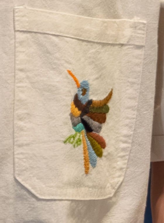 Extreme close-up of a chest pocket on a cream short-sleeve guayabera shirt with a hand-embroidered multicolor Otomi hummingbird in teal, orange, brown, grey, and olive green threads centered on the pocket flap