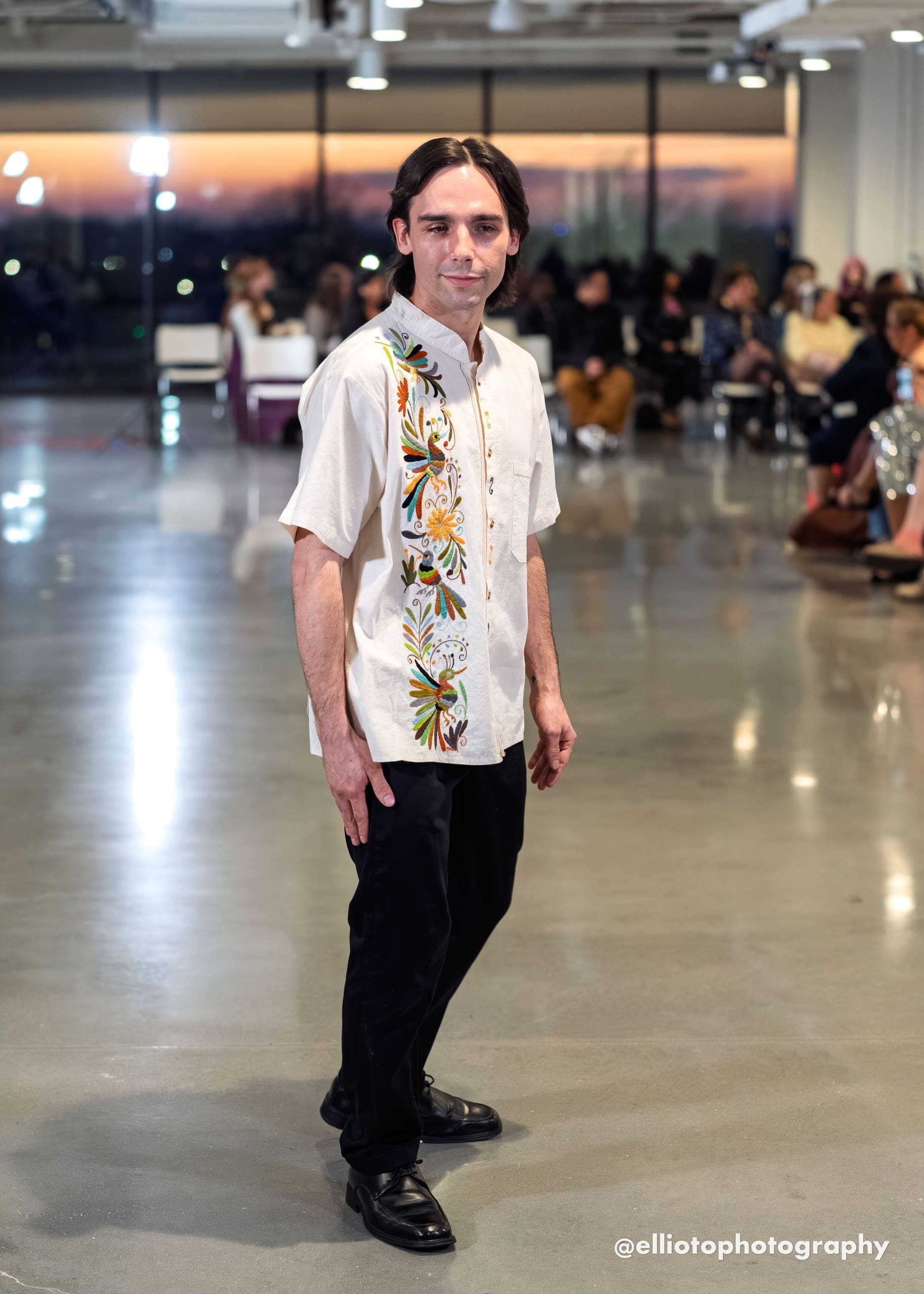 Male model walking a fashion runway wearing a cream short-sleeve mandarin-collar guayabera shirt with multicolor hand-embroidered Otomi hummingbirds, a golden daisy, and botanical motifs along the front placket, paired with black trousers and black oxford shoes