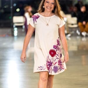 Smiling female model walking a fashion runway wearing a cream short-sleeve shift dress with hand-embroidered pink, magenta, and purple Otomi butterflies, daisy flowers, and botanical motifs on the lower front and left shoulder