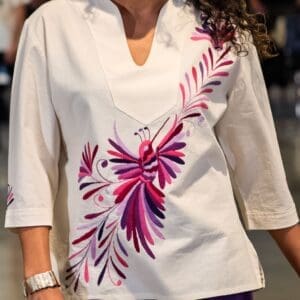 Front view close-up of a white v-neck blouse with large hand-embroidered pink, magenta, and purple Otomi hummingbird and feather motif worn by a model
