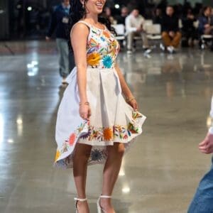 Smiling model twirling on a fashion runway in a white sleeveless fit-and-flare dress with multicolor hand-embroidered Otomi butterflies and flowers on the bodice and a floral border along the hem, worn with white platform heels