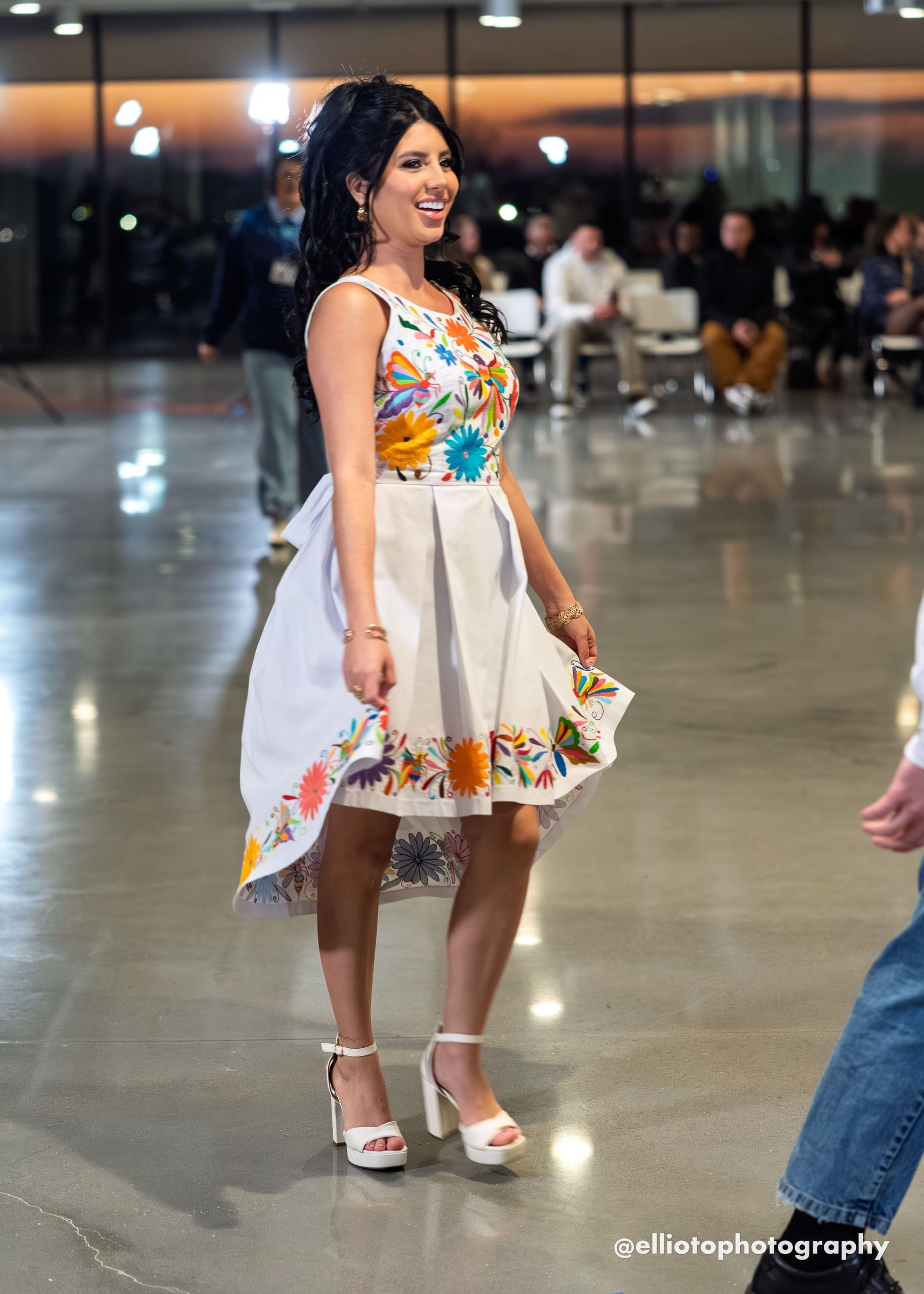 Smiling model twirling on a fashion runway in a white sleeveless fit-and-flare dress with multicolor hand-embroidered Otomi butterflies and flowers on the bodice and a floral border along the hem, worn with white platform heels