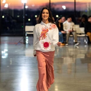 Female model walking a fashion runway wearing a white pullover hoodie sweatshirt with hand-embroidered pink, coral, and red Otomi bird and daisy flower motifs on the chest and kangaroo pocket, paired with a dusty rose wide-leg skirt.