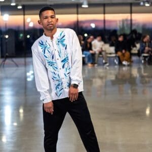 Male model walking a fashion runway wearing a white long-sleeve mandarin-collar guayabera shirt with hand-embroidered blue, teal, and grey Otomi dragonflies and botanical leaf motifs along the front placket, paired with black trousers and dress shoes