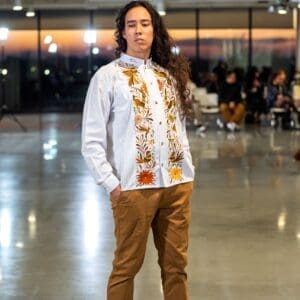 Female model walking a fashion runway wearing a white long-sleeve guayabera shirt with dual vertical bands of hand-embroidered earth-tone Otomi birds, rabbits, and daisy flowers in rust, olive, and brown, paired with tan chino trousers and black shoes
