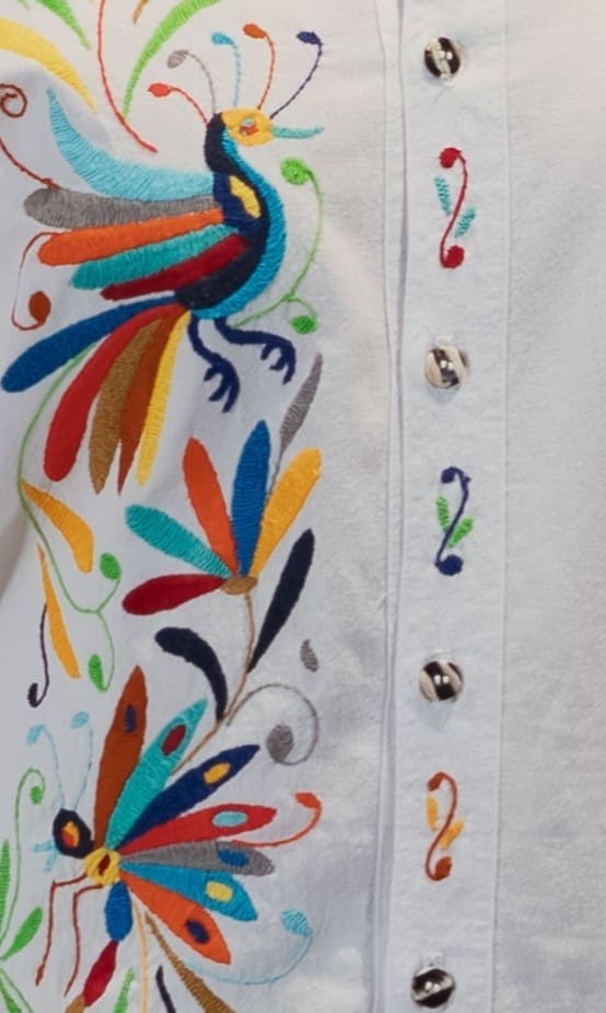 Close-up of the front placket of a white men's collared shirt showing hand-embroidered multicolor Otomi peacock with fanned tail, a large butterfly with teal and navy wings, and botanical leaf motifs in orange, blue, teal, yellow, and green on white cotton