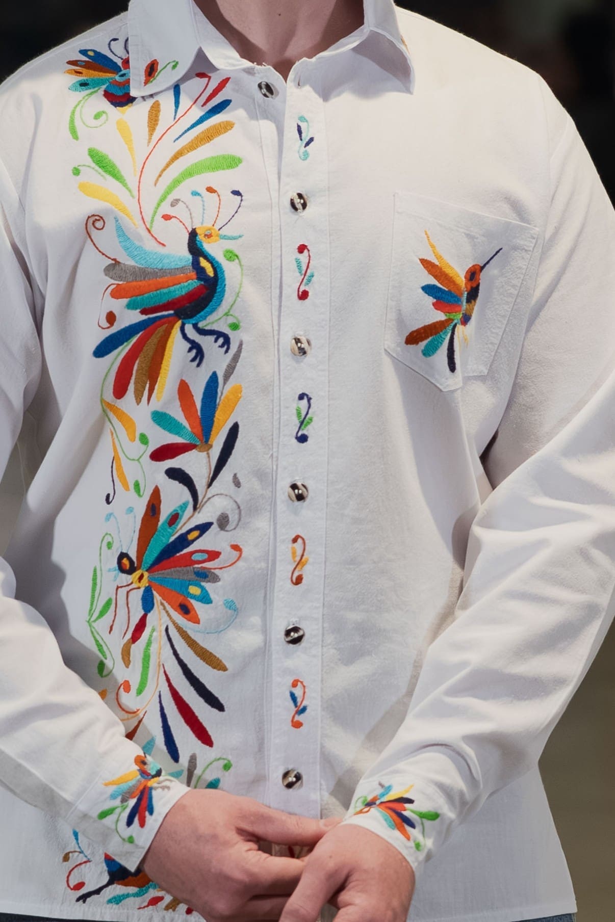 Close-up of a male model wearing a white long-sleeve collared button-down shirt with hand-embroidered multicolor Otomi peacock, butterfly, and botanical motifs along the front left placket in teal, red, orange, blue, green, and yellow, with a hummingbird on the chest pocket and embroidery on the cuffs