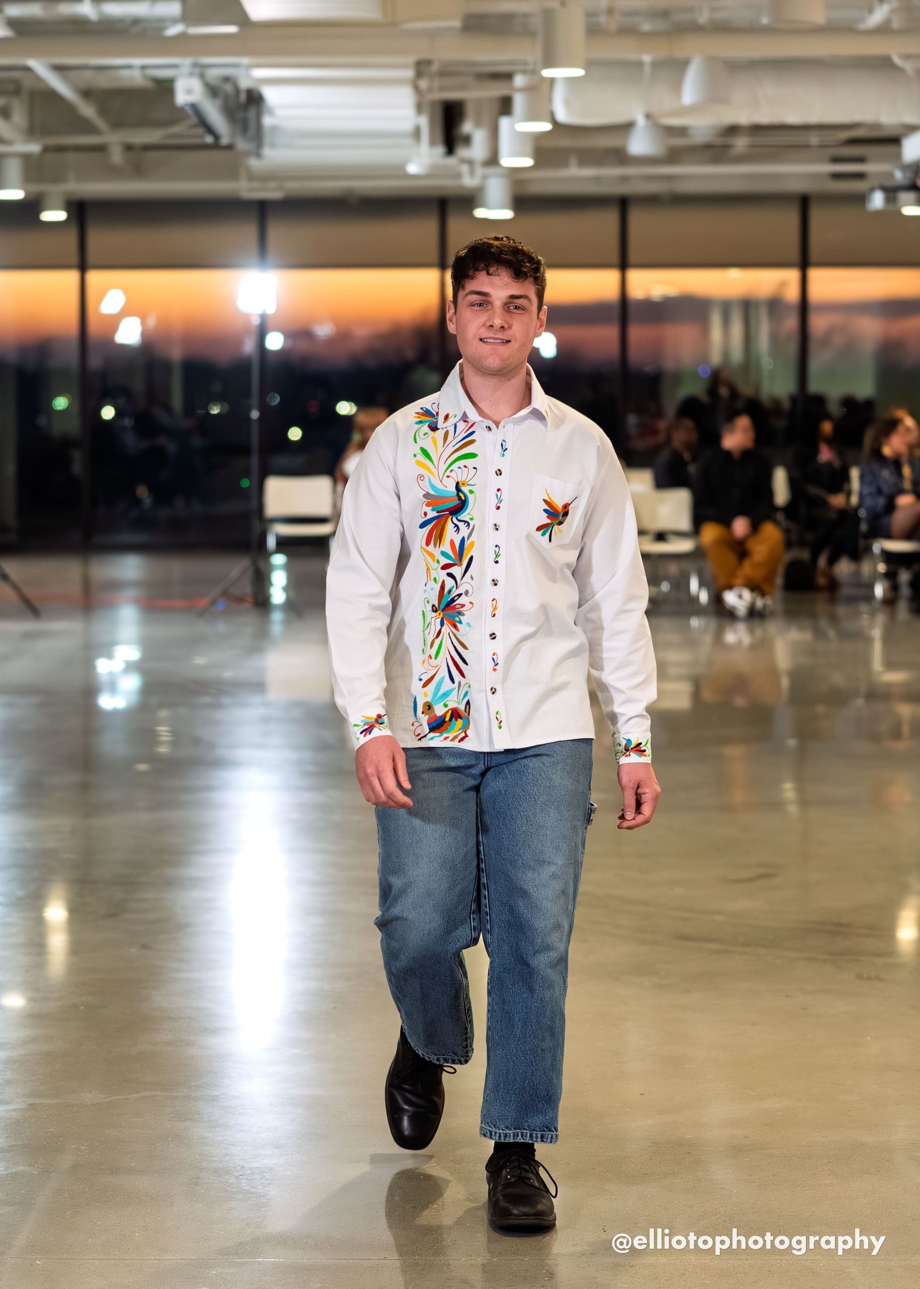 Male model walking a fashion runway wearing a white long-sleeve collared button-down shirt with multicolor hand-embroidered Otomi peacock, butterfly, and botanical motifs along the front left placket in teal, red, orange, blue, green, and yellow, paired with blue jeans and black dress shoes