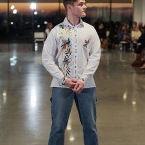 Male model on a fashion runway wearing a white long-sleeve button-down shirt with multicolor hand-embroidered Otomi peacocks, birds, and floral motifs in orange, blue, red, green, and yellow along the front placket and chest pocket, paired with blue jeans