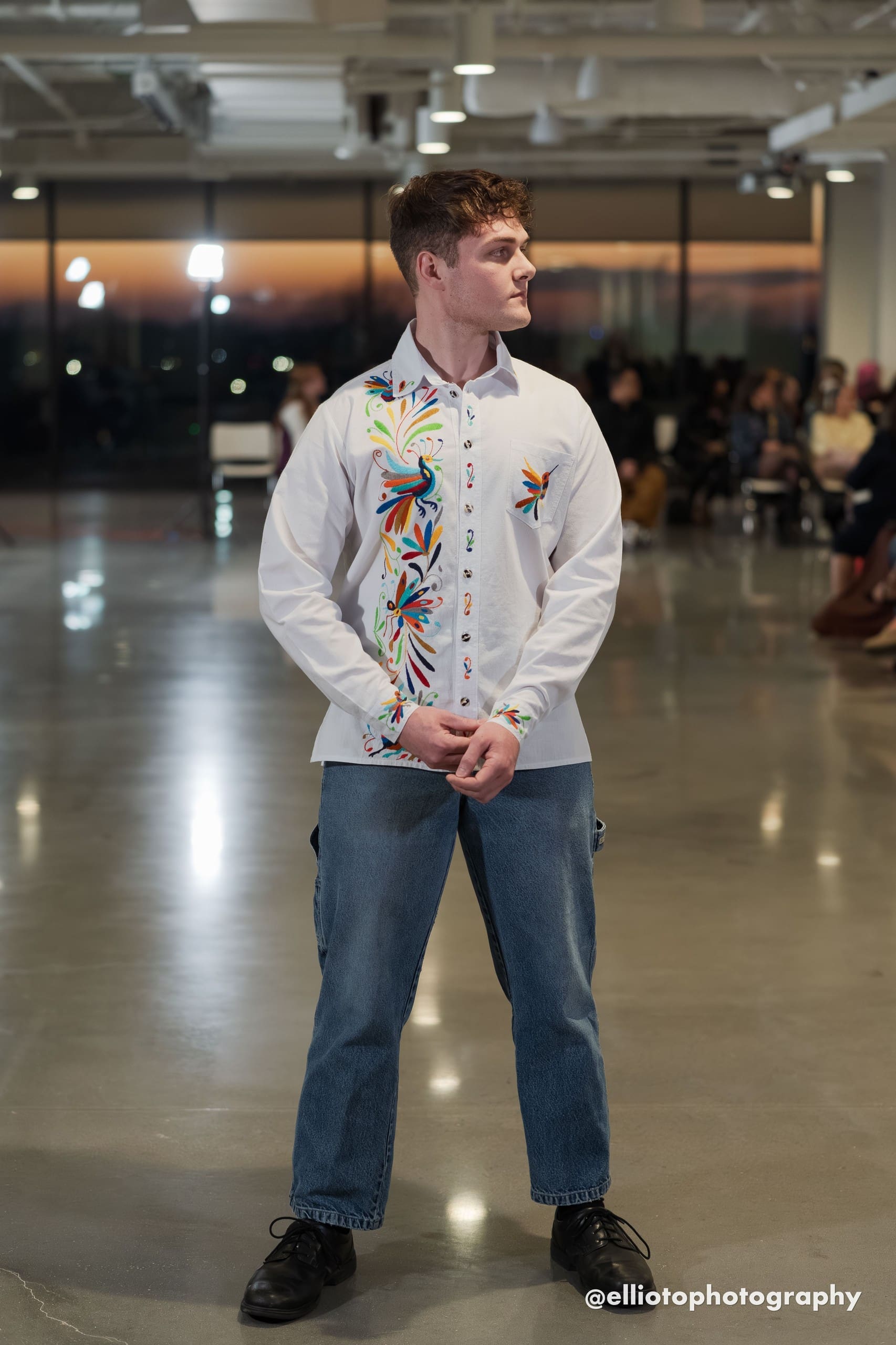 Male model on a fashion runway wearing a white long-sleeve button-down shirt with multicolor hand-embroidered Otomi peacocks, birds, and floral motifs in orange, blue, red, green, and yellow along the front placket and chest pocket, paired with blue jeans