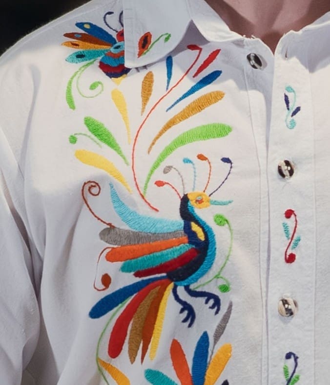 Close-up of the upper front placket of a white men's collared shirt showing a hand-embroidered multicolor Otomi peacock with fanned rainbow tail feathers in green, yellow, teal, red, orange, and grey, with botanical scrolls and a hummingbird visible near the collar