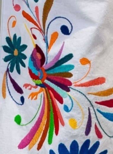 Close-up of multicolor hand-embroidered Otomi bird with fanned feather plumes and teal daisy flower on white cotton pants fabric, with botanical scrolls in orange, teal, and purple