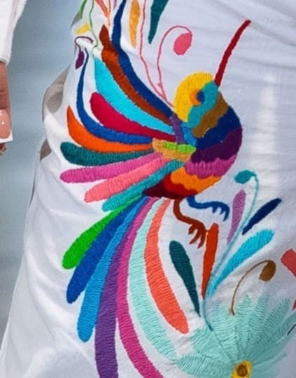 Close-up of a multicolor hand-embroidered Otomi hummingbird with rainbow feather plumes in teal, orange, pink, green, and blue on white cotton fabric