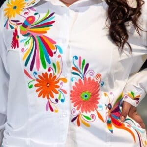 Woman wearing a white button-down cotton shirt with multicolor hand-embroidered Otomi birds, hummingbirds, daisy flowers, and butterflies in orange, pink, green, blue, and yellow threads