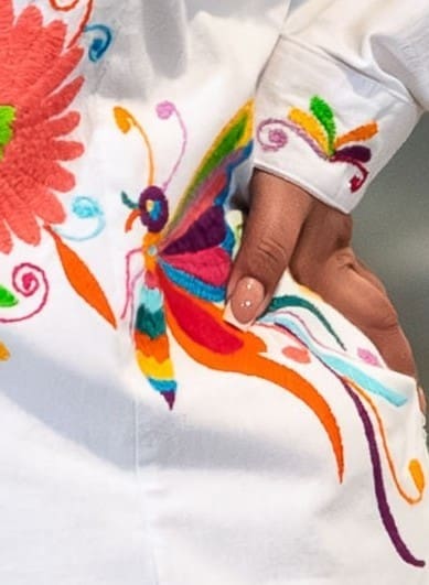 Close-up of a multicolor hand-embroidered Otomi butterfly and floral motif on the cuff and lower front of a white cotton shirt, with a model's hand visible