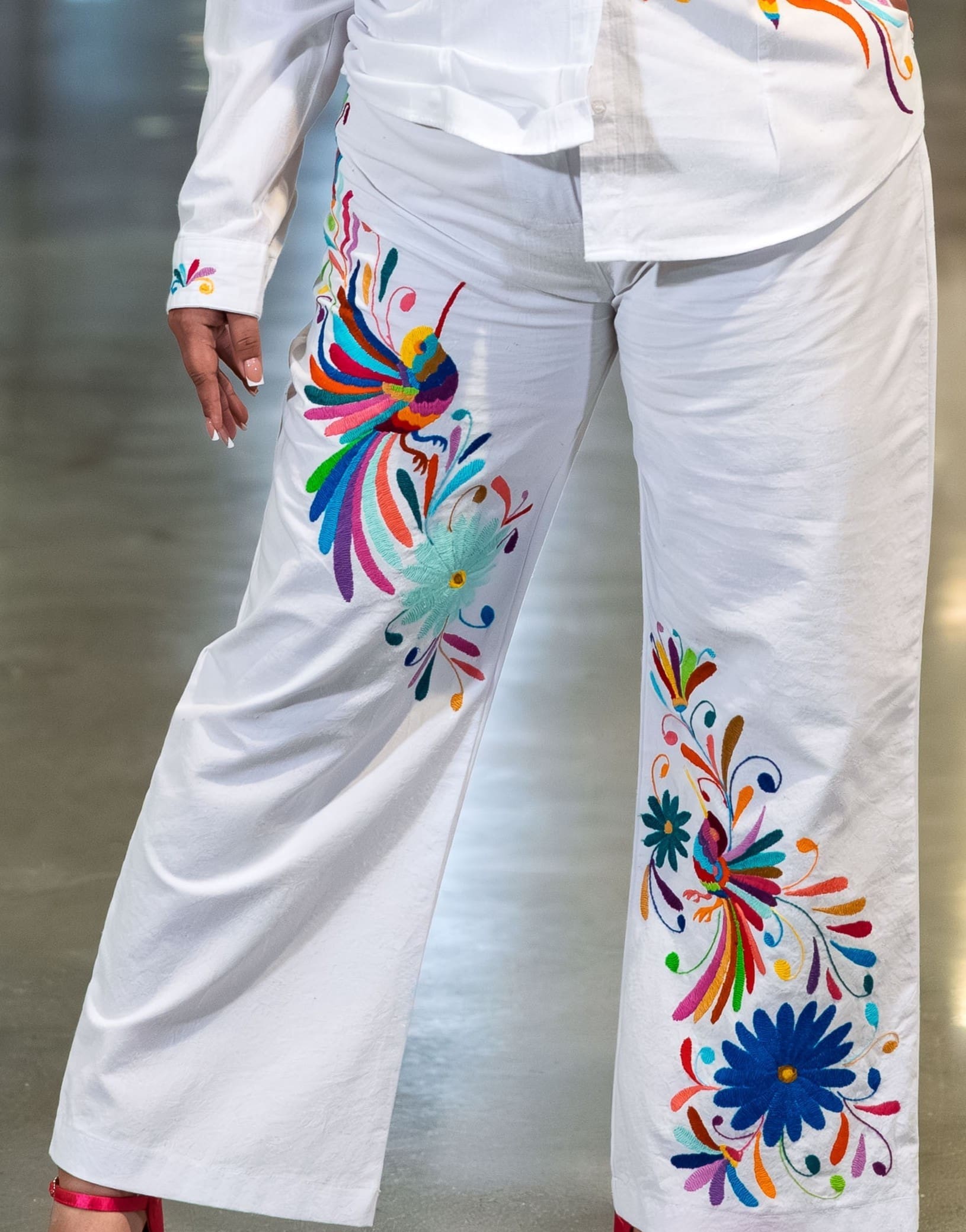 Close-up of a model wearing white wide-leg pants with multicolor hand-embroidered Otomi hummingbirds, daisy flowers, and botanical motifs in teal, orange, blue, pink, and green on both legs, paired with a white embroidered shirt
