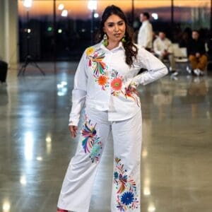 Female model on a fashion runway wearing white wide-leg pants with multicolor hand-embroidered Otomi birds, daisy flowers, and botanical motifs in orange, blue, red, green, and yellow, paired with a matching white embroidered shirt