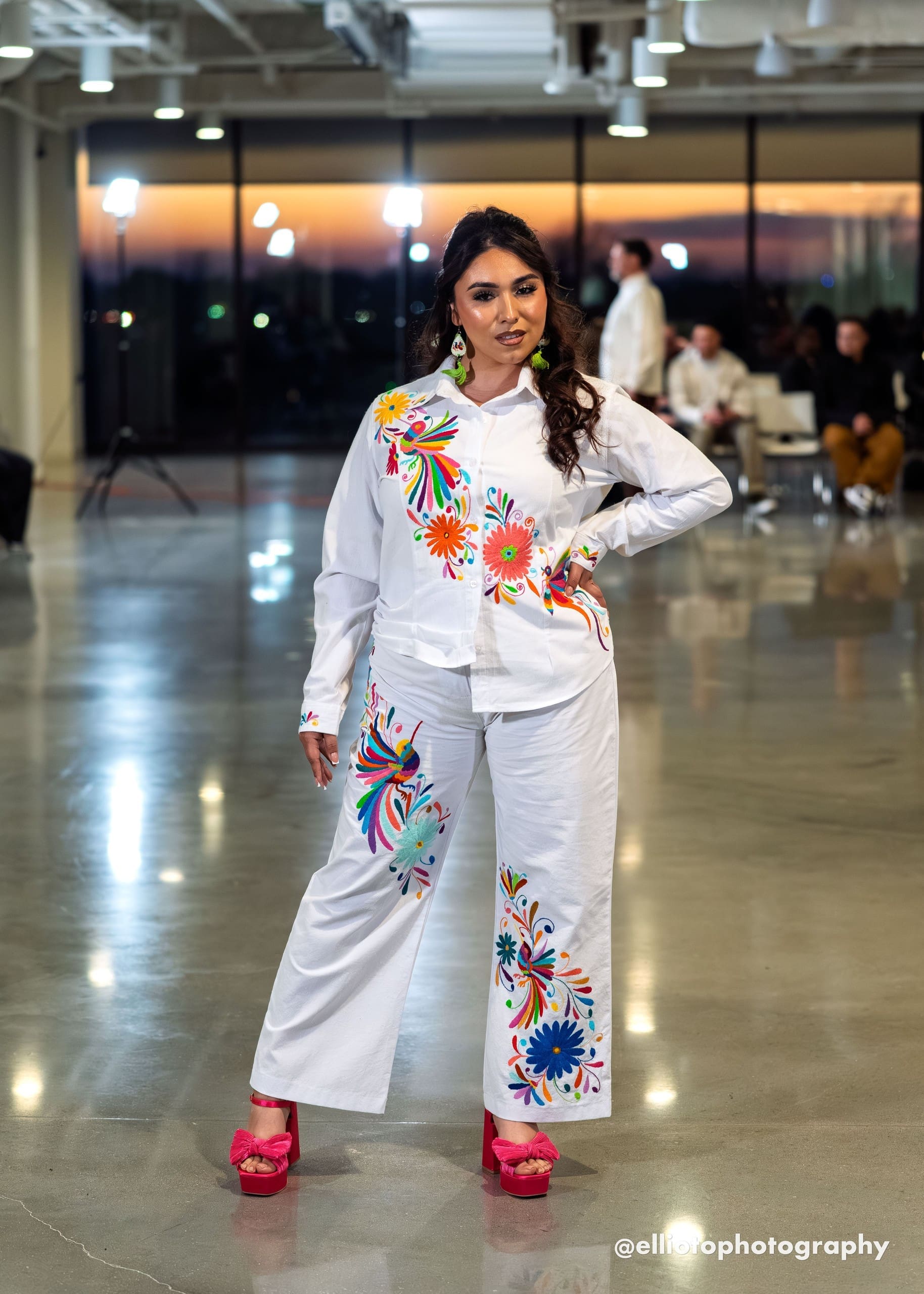 Female model on a fashion runway wearing white wide-leg pants with multicolor hand-embroidered Otomi birds, daisy flowers, and botanical motifs in orange, blue, red, green, and yellow, paired with a matching white embroidered shirt