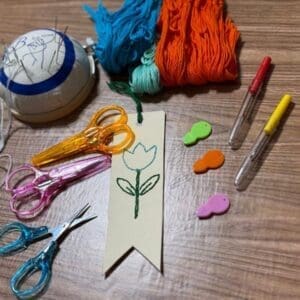 DIY embroidery bookmark kit featuring a hand-stitched green tulip, colorful skeins of thread, plastic needle threaders, seam rippers, and scissors on a wooden table for a B Magic Craft Hands workshop.