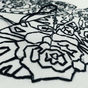 Close up of hand embroidery with a needle and black thread stitching a rose on white fabric in Bentonville, Arkansas. Acercamiento de bordado a mano con aguja e hilo negro delineando una rosa sobre tela blanca en Bentonville, Arkansas.